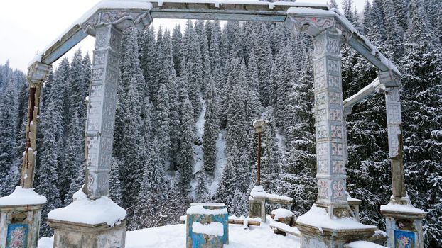 An old abandoned gazebo with views of the snowy mountains and forest. Clouds float beautifully, fir trees are covered with snow. High white hills. The stone pillars of the building are destroyed.