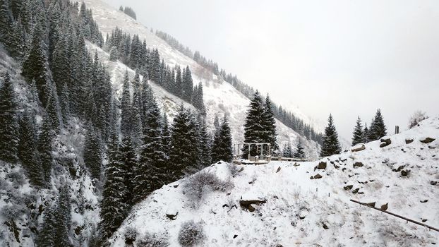 A broken gazebo stands on a snowy hill in the mountains. A beautiful gorge with tall fir trees and steep cliffs covered with snow. The river runs, it snows. People walk along gorge. Almarasan, Almaty