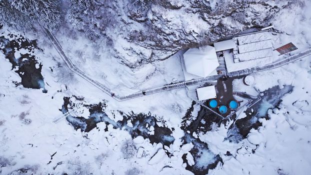 Hot springs in the snowy forest and mountains. Top view from the drone on the white gorge with the river. There are barrels of radon water. There is steam, people are bathing. Almarasan, Almaty.