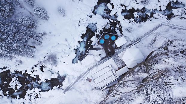 Hot springs in the snowy forest and mountains. Top view from the drone on the white gorge with the river. There are barrels of radon water. There is steam, people are bathing. Almarasan, Almaty.