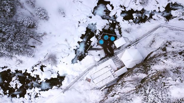 Hot springs in the snowy forest and mountains. Top view from the drone on the white gorge with the river. There are barrels of radon water. There is steam, people are bathing. Almarasan, Almaty.