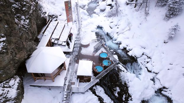 Hot springs in the snowy forest and mountains. Top view from the drone on the white gorge with the river. There are barrels of radon water. There is steam, people are bathing. Almarasan, Almaty.