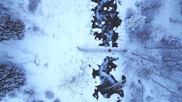 The guy is walking on the bridge over the river. The view from the drone to the tall trees, the river and the snowy gorge. The forest is covered with white snow. Mountainous terrain. Almarasan, Almaty