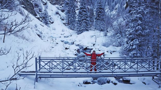 The guy is walking on the bridge over the river. The view from the drone to the tall trees, the river and the snowy gorge. The forest is covered with white snow. Mountainous terrain. Almarasan, Almaty