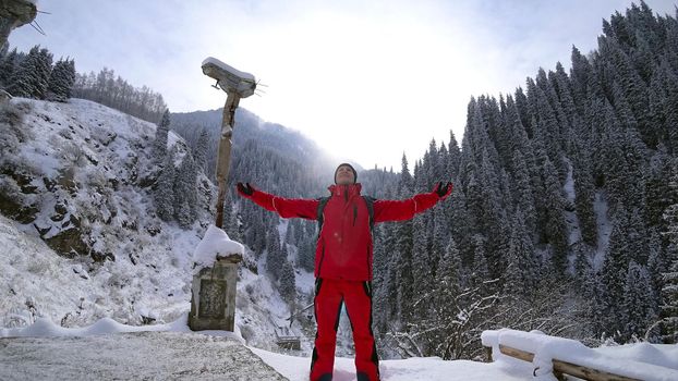 The guy stands on the edge of the hill and enjoys the view. Looks at a snowy gorge with high mountains, and fir trees growing on the cliffs. Clouds cover the mountain. Mountain suit with backpack.