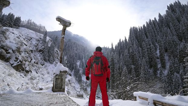 The guy stands on the edge of the hill and enjoys the view. Looks at a snowy gorge with high mountains, and fir trees growing on the cliffs. Clouds cover the mountain. Mountain suit with backpack.