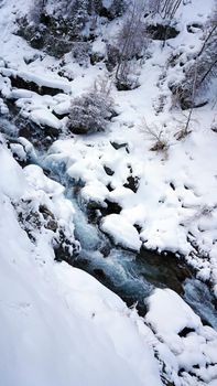 A clear mountain river runs through a snowy gorge. There is a metal bridge covered with snow. Tall spruce trees grow on the slopes of the mountains. There is steam from the river. Almarasan, Almaty