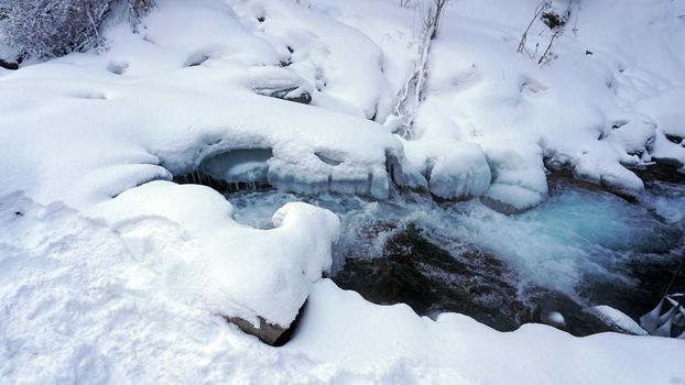 A clear mountain river runs through a snowy gorge. There is a metal bridge covered with snow. Tall spruce trees grow on the slopes of the mountains. There is steam from the river. Almarasan, Almaty