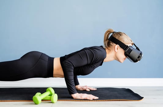 Fitness, sport and technology. Young athletic woman wearing virtual reality glasses doing push ups at blue background