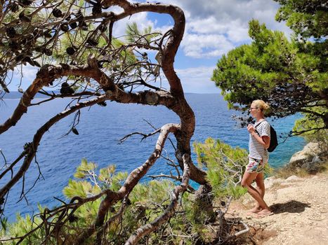 Young active feamle tourist wearing small backpack walking on coastal path among pine trees looking for remote cove to swim alone in peace on seaside in Croatia. Travel and adventure concept.