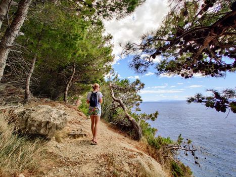 Young active feamle tourist wearing small backpack walking on coastal path among pine trees looking for remote cove to swim alone in peace on seaside in Croatia. Travel and adventure concept.