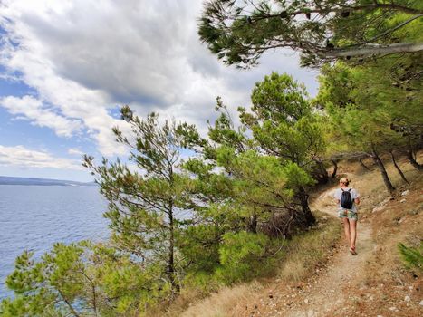 Young active feamle tourist wearing small backpack walking on coastal path among pine trees looking for remote cove to swim alone in peace on seaside in Croatia. Travel and adventure concept.