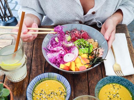 Woman eating tasty colorful healthy natural organic vegetarian Hawaiian poke bowl using asian chopsticks on rustic wooden table. Healthy natural organic eating concept.