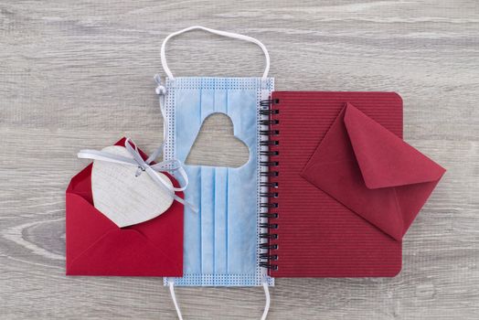 Face mask with cut out heart shape between a wire bound notepad and red envelope on wooden background with free copy space. Social distancing and contact-less greetings