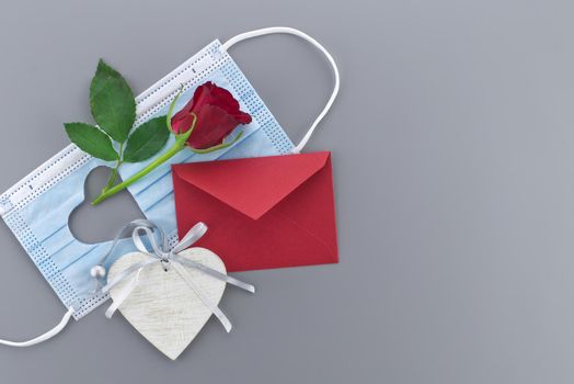 Medical mask with cut out heart shape, red rose envelope and wooden heart medallion on gray background with free copy space