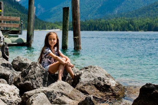 A little sweet girl sits on the shore of the Alpine lake. Plansee lake, Austria.