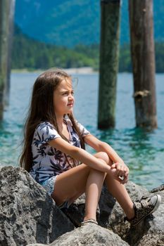 A little sweet girl sits on the shore of the Alpine lake. Plansee lake, Austria. Close-up.