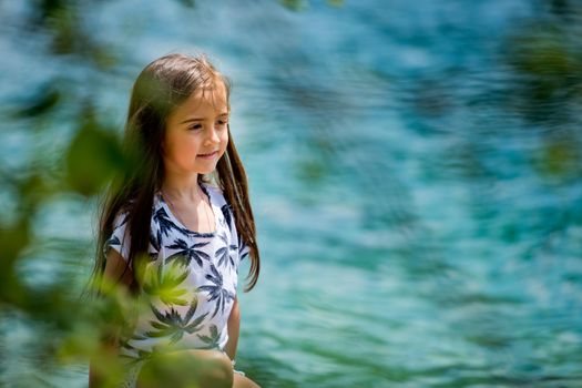 A little sweet girl sits on the shore of the Alpine lake. Plansee lake, Austria. Close-up.