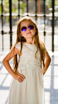 Portrait of a little girl in a white dress in sunglasses near the trellis fence. Close-up.