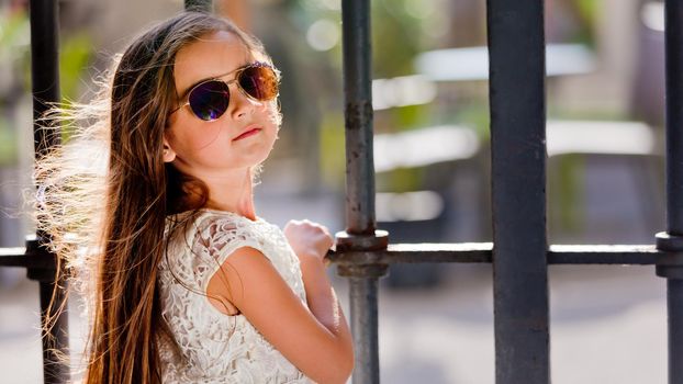 Portrait of a little girl in a white dress in sunglasses near the trellis fence. Close-up.