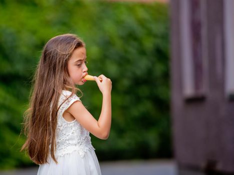 A little girl in a white dress is eating ice cream.