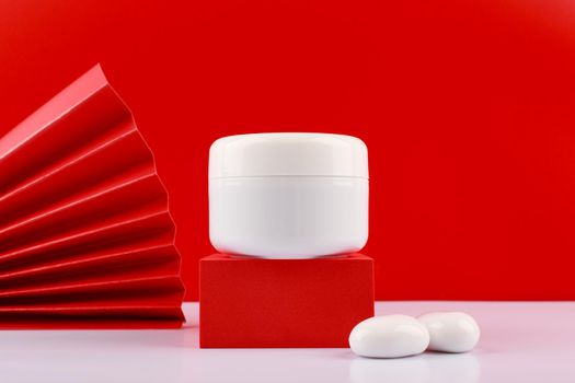 White cream jar on red podium with stones next to it against red background decorated with red waver. Concept of luxury skin or hair beauty product