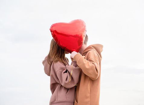 Valentines Day. Young loving couple hugging and holding red heart shaped balloon outdoors on sky background