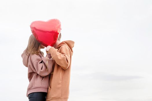 Valentines Day. Young loving couple hugging and holding red heart shaped balloon outdoors on sky background