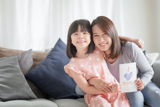 Asian mother hug her cute daughter that give handmade greeting card with i love mom word to surprising her at home