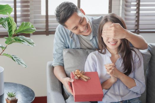 Handsome man is giving his girlfriend a red gift box for birthday surprising in the living room at home