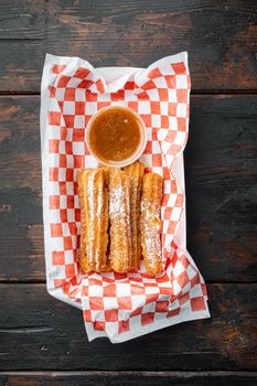 Tasty hot sweet churros in paper with chocolate, and caramel in paper tray, on old dark wooden table background, top view flat lay