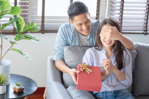 Handsome man is giving his girlfriend a red gift box for birthday surprising in the living room at home