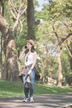 Beautiful asian solo tourist woman  smiling and enjoy taking via mobile phone while walking in public nature park. Vacation travel in summer.