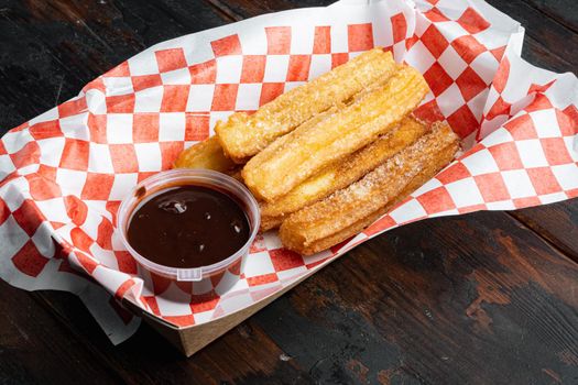 Tasty hot sweet churros in paper with chocolate, and caramel in paper tray, on old dark wooden table background