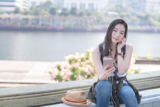 Beautiful asian solo tourist woman relaxing and enjoying listening the music on a smartphone in urban city downtown. Vacation travel in summer.