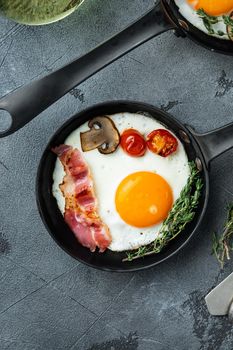Fried eggs with bacon and vegetables set in cast iron frying pan, on gray background, top view flat lay