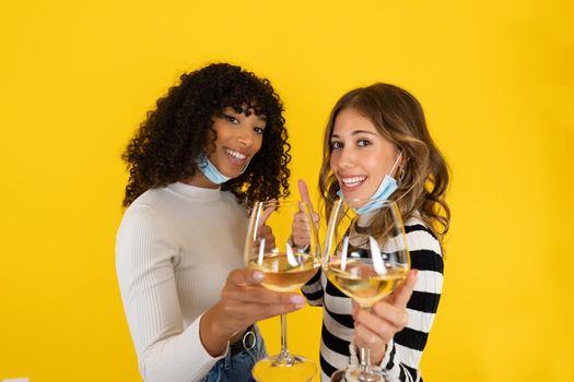 Two young women isolated on yellow background doing thumb up sign wearing protection mask holding white wine glass and smiling looking at the camera. Optimist concept of celebrate end of pandemic