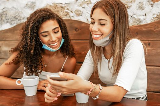 Two girls wearing protection mask sitting at bar table looking smartphone with coffee cups. New normal human habits: having fun with social network technology and healthy care. Focus on right woman