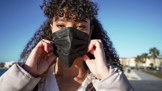 Close up portrait of young African American woman while wearing a black mask to avoid Coronavirus infection. Outdoor new normal social behaviors for human in the city against pandemic illness.