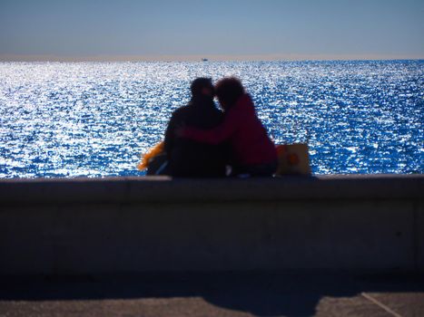 Blurred image of unrecognizable embraced lovers seen from behind sitting in front of the sea