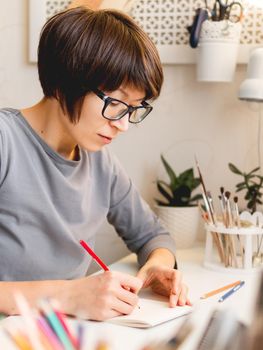 Woman with short hair cut is drawing in notebook. Calming hobby, anti stress leisure. Artist at work. Cozy workplace.