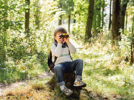 Little explorer on hike in forest. Boy with binoculars sits on stump. Outdoor leisure activity for children. Summer journey for young tourist.