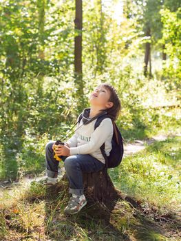 Little explorer on hike in forest. Boy with binoculars sits on stump. Outdoor leisure activity for children. Summer journey for young tourist.