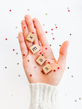 Word XMAS on palm of hand. Woman in white knitted sweater holds wooden letters with stars confetti. Christmas and New Year background.