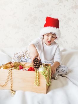 Joyful boy in Santa Claus hat is playing with decorations for Christmas tree. Funny kid is ready for New Year celebration. Cozy home. Winter holiday spirit.