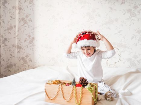 Joyful boy in Santa Claus hat is playing with decorations for Christmas tree. Funny kid is ready for New Year celebration. Cozy home. Winter holiday spirit.