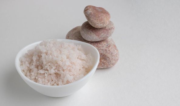 Pink coarse salt in a white bowl stands on a white background next to pink stones.