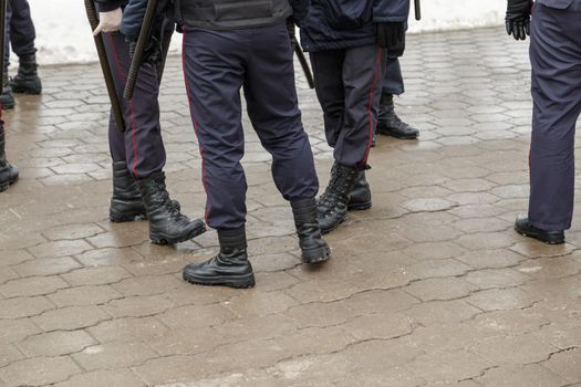 Russian police officers legs in black ankle boots, red stripe pants and with rubber tonfa night sticks on wet pavement surface - close-up with selective focus and blur.