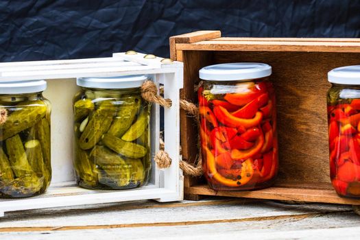 Wooden crate with glass jars with pickled red bell peppers and pickled cucumbers (pickles) isolated. Jars with variety of pickled vegetables. Preserved food concept in a rustic composition.