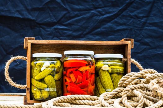 Wooden crate with glass jars with pickled red bell peppers and pickled cucumbers (pickles) isolated. Jars with variety of pickled vegetables. Preserved food concept in a rustic composition.